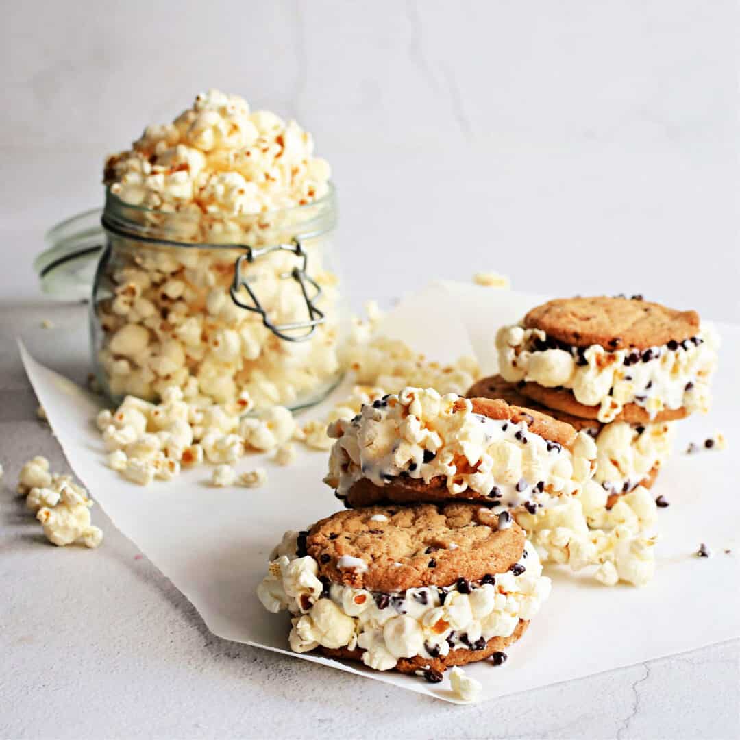Popcorn ice cream sandwiches combine chocolate chip cookies, vanilla ice cream and crunchy popcorn into one amazing sweet treat. Ice cream sandwiches themselves date back to the start of the 20th century so we all grew up with this frozen summer treat. Two soft, thin, chocolate cake layers with a slab of vanilla ice cream in the middle and they're still very much around, both in grocery stores and ice cream trucks. And they're still just as good. But then someone came up with the idea of sandwiching the ice cream between two cookies and we applaud them. The famous Chipwich can be found everywhere and, not only does it use chocolate chip cookies but the outside edge is rolled in more chocolate chips. It's not hard to make your own version and it gives us the opportunity to make the recipe even more interesting. We kept the cookies, kept the chocolate chips, kept the vanilla ice cream but added popcorn. Yup, we roll the softened ice cream edge in popcorn kernels for an extra crunch and that popcorn flavor we love so much. Rest assured that we've put these poporn-ized desserts in the freezer to make sure they don't go soggy. Nope. They actually hold up great. As much as we love this recipe with a standard chocolate chip cookie, albeit with mini chips, we also think it would be amazing with our salted caramel popcorn cookies. They are sweet, salty, tender and crunchy with popcorn baked right into the batter and dotted on the outside. Thanks to our evil genius experiments, we know that the popcorn does not become mushy. It maintains enough texture to be fun to eat. You can find our recipe HERE or use the chocolate chip cookie recipe below. Note that our popcorn cookies are slightly larger than the ones in this recipe so just use a smaller scoop. Or, don't. Make 'em big. Add more ice cream. We're not judging you.